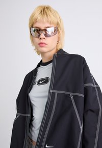 Casaco oversized preto com costura branca sobre uma t-shirt cinza com um logo preto. O modelo usa óculos de sol refletivos.