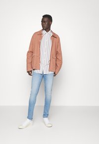 Chaqueta ligera de color rosa sobre una camiseta de rayas blanca y gris, combinada con jeans de color azul claro y zapatillas blancas. Texturas suaves, corte relajado.