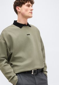 Νέος άνδρας που φοράει ελιάς πράσινη φούτερ BOSS πάνω από μαύρο πουκάμισο με γιακά, γκρι παντελόνια και μαύρη ζώνη με ασημένια αγκράφα.