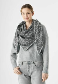 Femme portant un pull gris et un jean, avec une grande écharpe à motif léopard gris enroulée autour du cou, souriant légèrement.