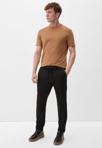 Bruine T-shirt met korte mouwen en ronde hals, gecombineerd met zwarte joggingbroek met trekkoord en zwarte sneakers met gestructureerde zolen, staand op een witte achtergrond.