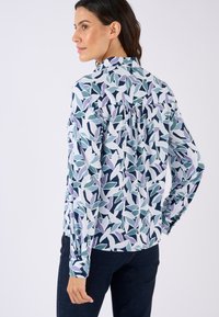 Langarmshirt in een bloemenpatroon met groene, paarse en witte bladeren op een lichtblauwe achtergrond; gare knopen bij de mouwen en plooidetail op de rug.