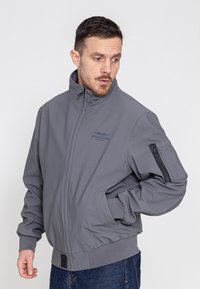 Grau Bomberjacke mit hohem Kragen, Reißverschlusstasche am Ärmel, gerippten Bündchen und Saum, aus glattem Stoff gefertigt. "BOMBERS"-Logo auf der Brust.