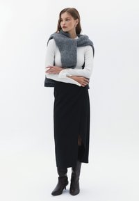 Falda negra envolvente con una abertura lateral, combinada con una blusa blanca de manga larga y textura, y una bufanda de punto gris colocada sobre los hombros. Botas de tacón marrones.