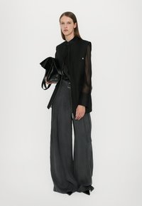 Melnā veste ar caurspīdīgām piedurknēm, kombinēta ar melnu blūzi un brīviem pelēkiem biksēm. Modelis tur nelielu melnu somiņu. Balta fonā.