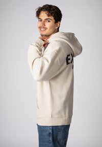 Beiger Hoodie mit lockerem Schnitt, Kängurutasche und gerippten Bündchen. Auf der Rückseite schwarzer Text. Kombiniert mit blauer Jeans.