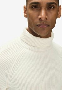 Valkoinen ribattupintainen turtleneck-paita, jossa on teksturoitu kangas. Lähikuva, joka näyttää pehmeän materiaalin ja istuvan muotoilun, korostaen kaulusta ja ompeluyksityiskohtia.