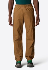 Pantalons légers marron avec une taille élastique, jambes fuselées et détail de logo discret. Associés à des chaussures grises avec des accents verts.