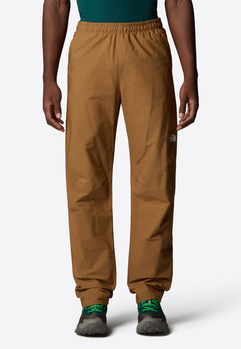 Pantalons légers marron avec une taille élastique, jambes fuselées et détail de logo discret. Associés à des chaussures grises avec des accents verts.