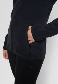 Tummansininen fleece-takki, jossa on vetoketju, sivutaskut ja sileä pinta, päällä mustat leggingsit. Näkyvät käsi taskussa -tiedot.