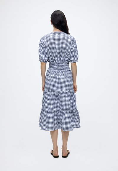 Femme debout de dos portant une robe à carreaux bleue et blanche avec des manches bouffantes et une jupe midi à volants, sandales plates noires.
