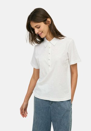 Mujer con cabello castaño que lleva una camisa blanca de manga corta abotonada y jeans azules, mirando hacia abajo con una ligera sonrisa.