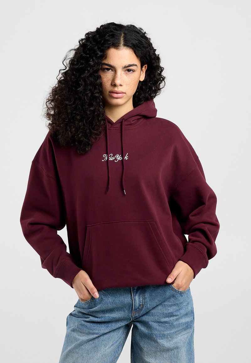Mujer joven con cabello rizado que lleva una sudadera con capucha burdeos con el texto "New York", manos en el bolsillo frontal, combinada con jeans azules sobre un fondo liso.