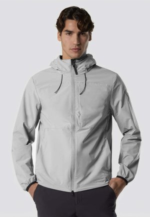 Giovane uomo che indossa una giacca grigia chiara con cappuccio e zip frontale e pantaloni neri, in piedi con le mani in tasca su uno sfondo chiaro.