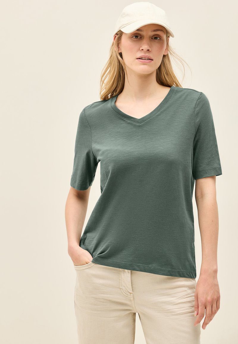 Oliven grøn kortærmet V-hals t-shirt lavet af blødt materiale. Har en afslappet pasform og er parret med lysebeige bukser.