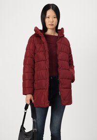 Doudoune rouge avec capuche, présentant un design matelassé et des boutons-pression. Portée sur un pull sombre et associée à un jean foncé et un sac noir.