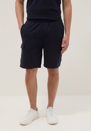 Mann trägt schwarze Cargo-Shorts mit Seitentaschen, schwarzes Hemd und weiße Sneakers, steht mit den Händen in den Taschen vor einfachem Hintergrund.