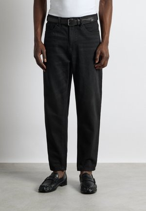 Mand iført sorte jeans med sort bælte, hvid skjorte og sorte læder loafers, står på en grå gulv mod en hvid væg.