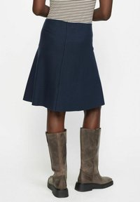 Jupe évasée bleu marine en tissu extensible, avec une texture lisse et une silhouette longueur genou, associée à des bottes marron jusqu'aux genoux.
