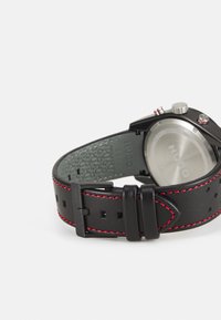 Black leather watch strap with red stitching, metal hardware, and textured backing. Round silver watch face, brand logo on the back.