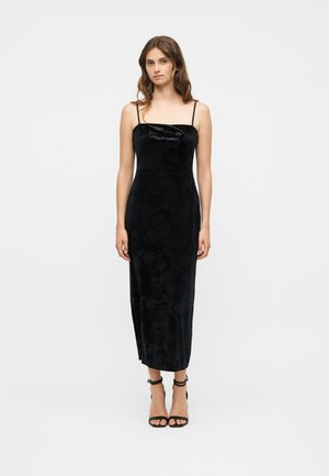 Mujer de pie, vestida con un vestido midi sin mangas de terciopelo negro con tirantes finos y tacones altos negros con correa en el tobillo sobre un fondo blanco liso.