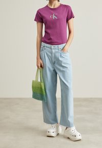 Fialové bavlněné tričko s logem "ck", světle modré široké džíny, bílé dřeváky a kabelka ze síťoviny s zeleným gradientem.