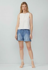 Top sans manches blanc avec des détails en œillets, associé à un short en denim bleu clair avec une taille à cordon de serrage et des ourlets roulés. Sandales beiges à lacets.