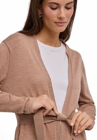 Beige cardigan met een zachte textuur, open voorkant en lange mouwen. Voorzien van een ceintuur om zelf te knopen. Draagt over een wit overhemd.