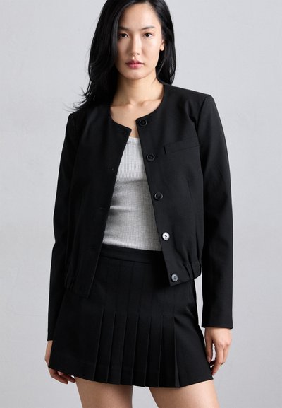 Svart jakke med rund hals og trykknapper, kombinert med et svart plissert skjørt. Stoffet ser glatt ut; designet er minimalistisk med rene linjer.