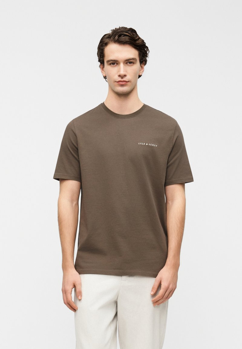 Καφέ βαμβακερό t-shirt με κοντά μανίκια, στρογγυλή λαιμόκοψη και λευκό λογότυπο στο στήθος. Λεία υφή και άνετη εφαρμογή, συνδυασμένο με ανοιχτόχρωμο παντελόνι.