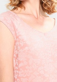 Kvinna i en ljusrosa blommig spets-topp med korta ärmar och en rundad kvadratisk halsringning.