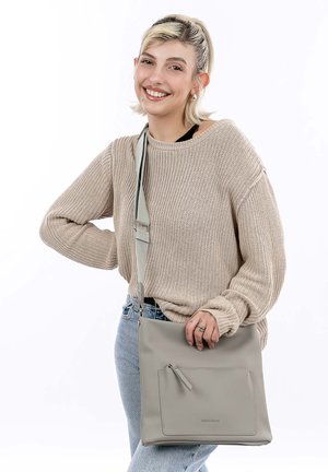 Young person with short blonde hair smiles, wearing beige sweater and light blue jeans, holding gray crossbody bag with front pocket.