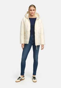 Puffy cream jacket with hood, front zipper, and drawstrings; paired with dark blue top and skinny jeans; finished with black and white sneakers.