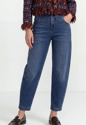 Person iført mørkeblå jeans, en tekstureret brun og orange jakke og mørkebrune læder loafers, stående med den ene hånd i lommen.