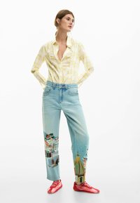 Cămașă în carouri galbenă cu guler deschis, blugi din denim bleu deschis cu grafice colorate reprezentând un peisaj de coastă și pantofi sport roșii.