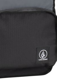 Mochila negra con base y parte superior gris, que presenta una tela texturizada, un bolsillo y un parche rectangular con el logo de Volcom en la parte frontal.