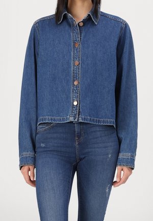 Vrouw met een blauwe denimjas met knopen en bijpassende blauwe jeans met lichte slijtage op de dijen, staande tegen een effen achtergrond.