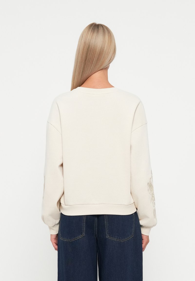 Kremowy sweatshirt z obniżonymi ramionami, prążkowanymi mankietami i haftowanym detalem na lewym rękawie. W połączeniu z ciemnoniebieskimi spodniami z szerokimi nogawkami.