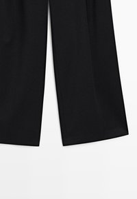 Pantalons larges noirs avec une fente centrale à l'avant de chaque jambe, présentés sur un fond blanc uni.