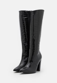 Bottes noires montantes en cuir verni avec une texture crocodile, bout pointu et talon chunky, dotées d'une fermeture éclair à l'arrière.