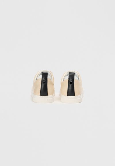 Sneakers din suede bej, cu o dunga texturată neagră la toc, talpă albă și un design minimalist. Literele clare ale brandului pe spate.