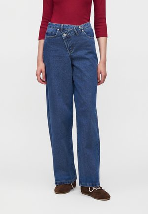 Person iført blå højttaljede jeans med en asymmetrisk knapfastgjort linning, kombineret med brune sko og en rød langærmet top.