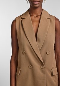 Hnědý bezrukávový blazer s hlubokým výstřihem do V, dvouřadým zapínáním, dvěma knoflíkovými detaily a bočními kapsami z texturované látky.