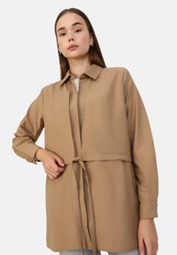 Light brown, long-sleeve blouse made of smooth fabric. Features a collar, front buttons, and a tie at the waist for adjustable fit.