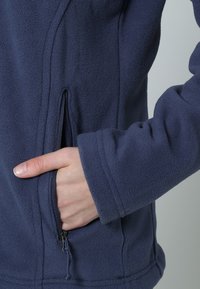 Merinovärinen fleece-takki, jossa on pehmeä pinta, varustettuna vetoketjullisella taskulla ja sileällä kauluksella. Hihansuut ovat istuvat ja hieman levenevät.