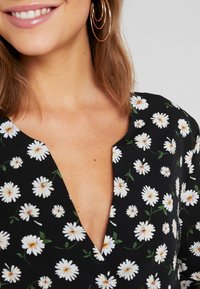 Robe noire à fleurs avec des marguerites blanches, design à encolure en V, tissu lisse, présentant un gros plan du motif floral et du détail de l'encolure.