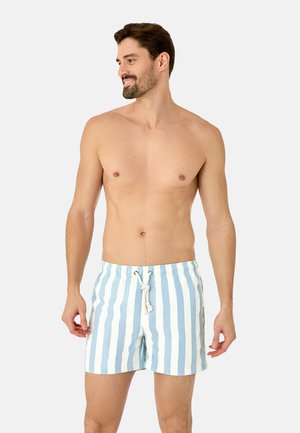 Mand smiler og kigger til siden, iført lyseblå og hvide lodrette stribede badeshorts med snor på en lys baggrund.