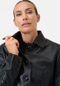 Zwarte leren jas met een gesneden kraag, voorzien van grote zwarte knopen en een gladde textuur, met een close-up opname van een model.