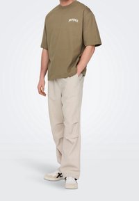 Oliwkowa bawełniana koszulka z grafiką "MONET", w połączeniu z jasnobeżowymi plisowanymi spodniami i białymi trampkami z czarnymi akcentami.