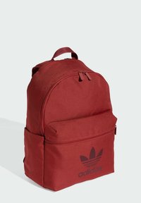 Zaino in tessuto rosso con forma tondeggiante, manico superiore, tasca frontale con zip e logo Adidas nero sul davanti in basso. Texture liscia.